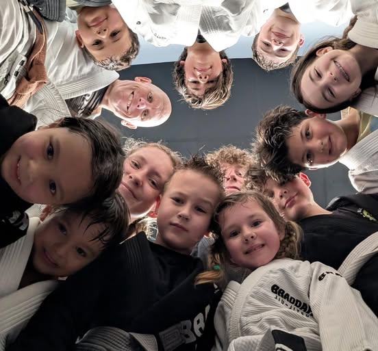 Braddah Jiu Jitsu children and adults in white and black gis gathered together and looking down at the camera during a group training session.